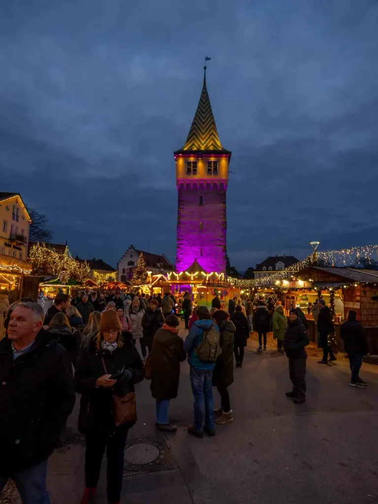 A Christmas market with illuminated stalls, many visitors and a pink illuminated tower at Lindau am Bodensee