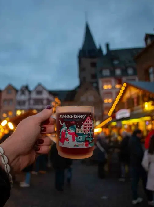 Hand holding a Christmas mug in front of a Christmas market with lights surrounded by houses and a church